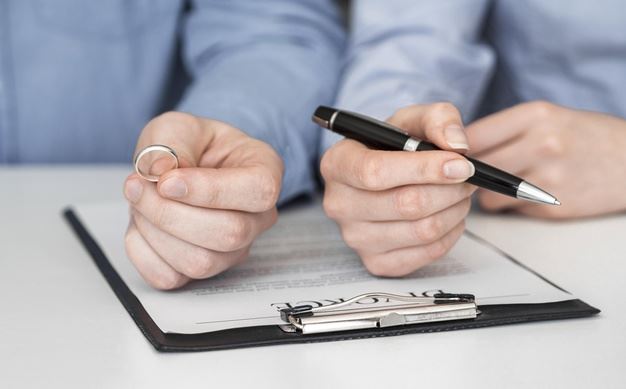Close up of one hand holding a ring and another hand holding a pen over a divorce contract.